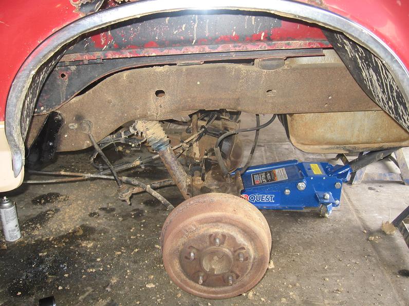Red vehicle rear wheel well with wheel off, showing rusty brake drum and suspension, blue floor jack underneath