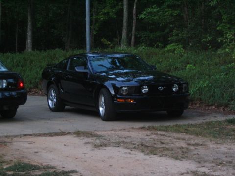 2007 Ford Mustang - A black Ford Mustang is parked on a dirt and concrete surface with trees in the background.