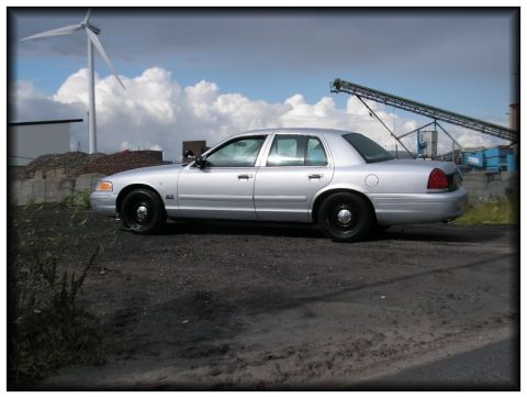 1998 Ford Crown Victoria - Side view of a silver 1998 Ford Crown Victoria parked outdoors near industrial structures.