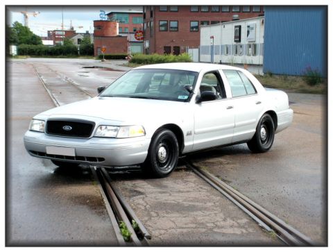 1998 Ford Crown Victoria - A white sedan is parked on train tracks in an urban setting.