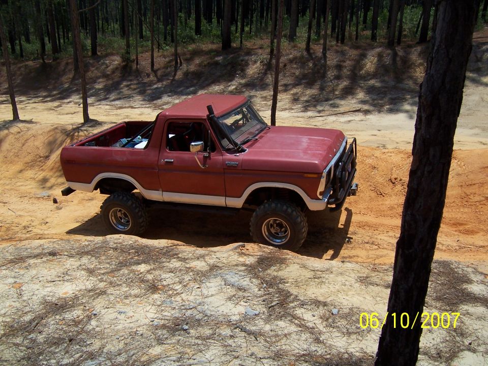 Topless red Ford Bronco with roll bar and mud tires parked on orange sand in a pine forest