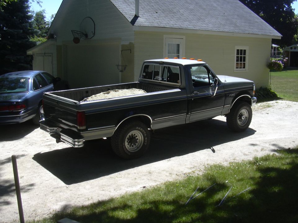 Dark blue and gray Ford F-250 pickup truck parked in a driveway with sand in the bed. A blue sedan is parked to its left