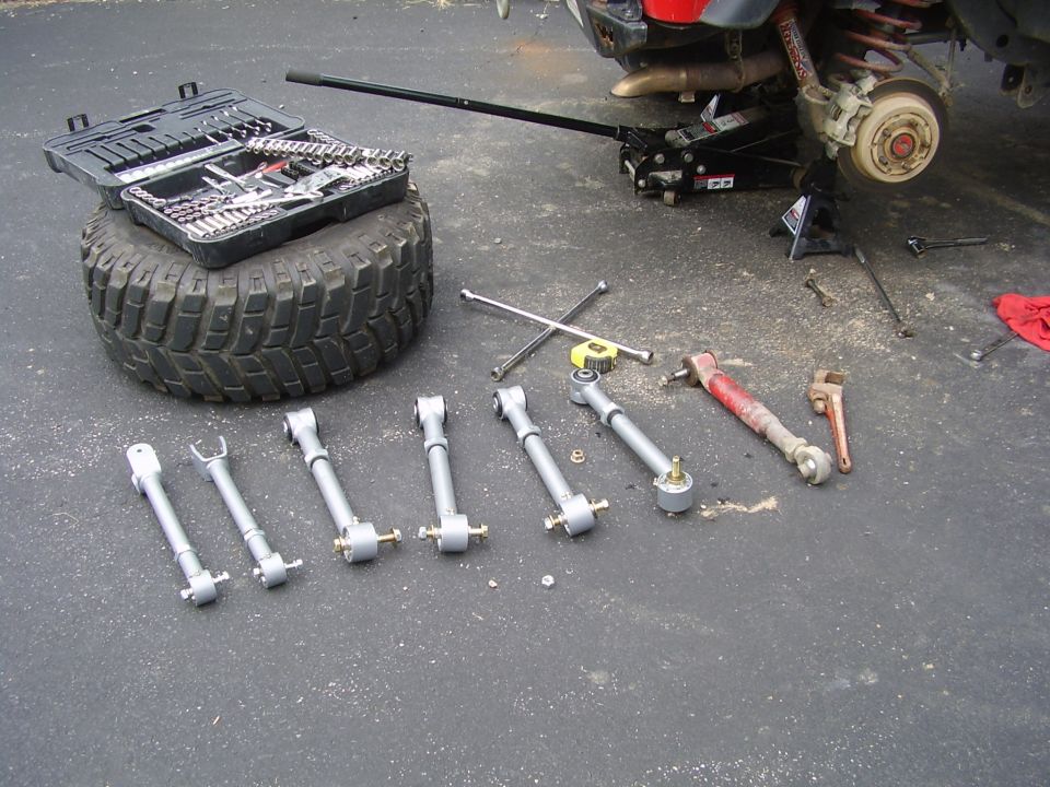 Tire, toolbox, suspension control arms, jack, and wrenches laid out on asphalt