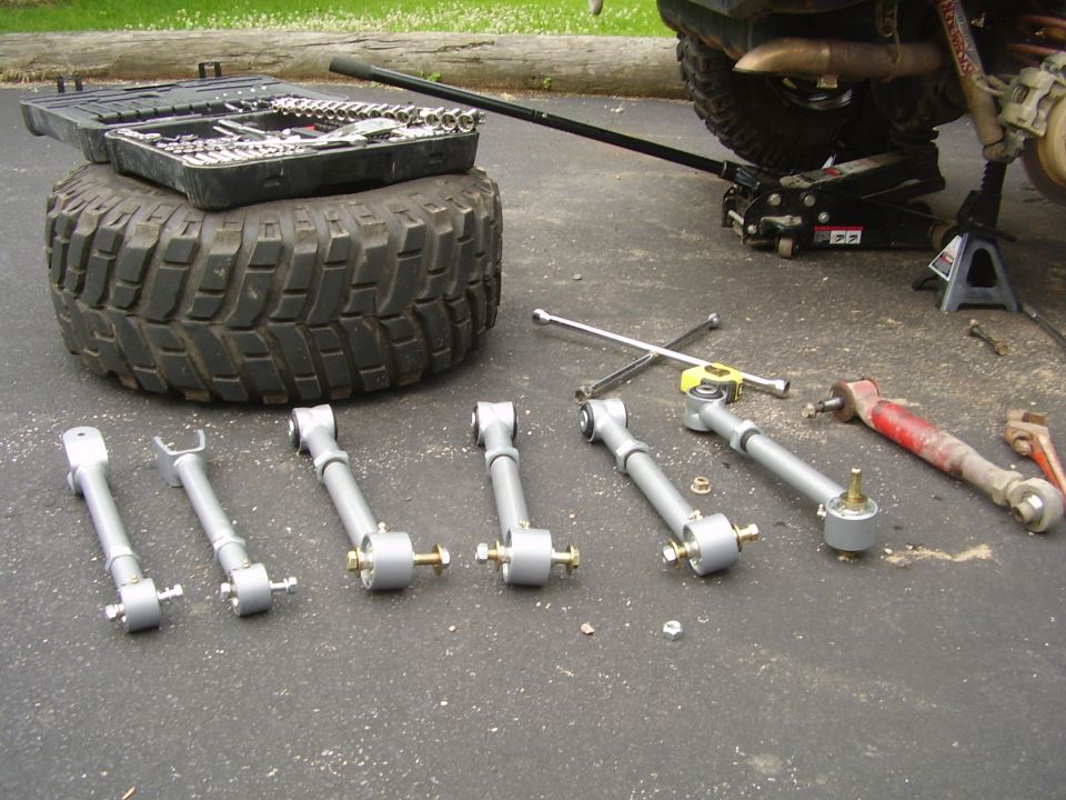 Several silver suspension control arms laid out on asphalt next to a tire, toolbox, and jack
