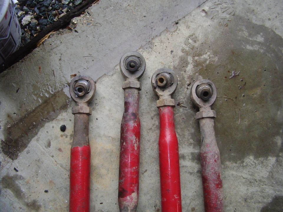 Four red suspension control arms with metal ends, some showing rust and dirt, on a concrete surface