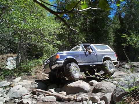 1989 Ford Bronco - A blue Ford Bronco tackles a challenging rocky terrain on a wooded trail.