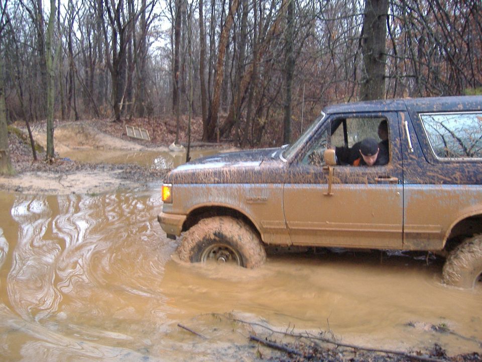 Ford Bronco stuck in a muddy puddle in a wooded area. Driver is visible inside