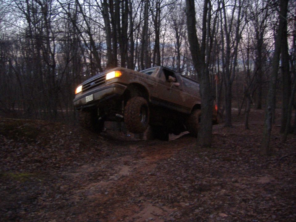 Brown Ford Bronco climbing a muddy hill in a forest, front right wheel lifted
