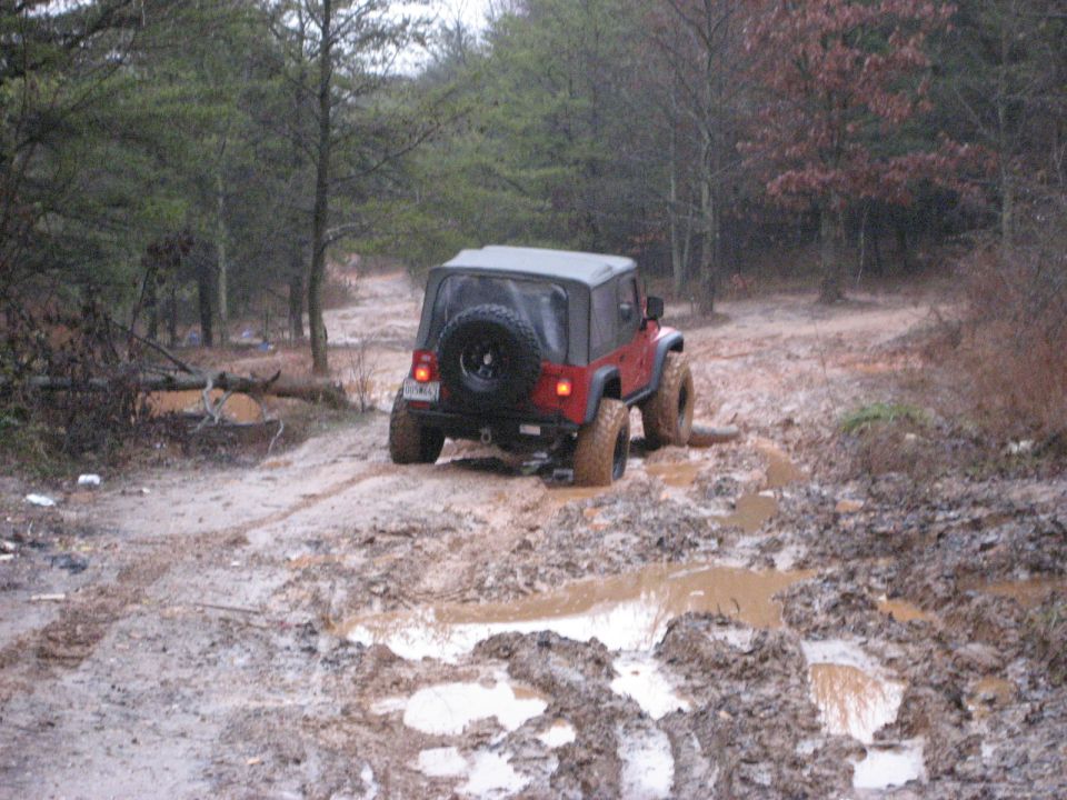 Red Jeep Wrangler driving through a muddy trail in the woods. It's got a soft top and a spare tire on the back
