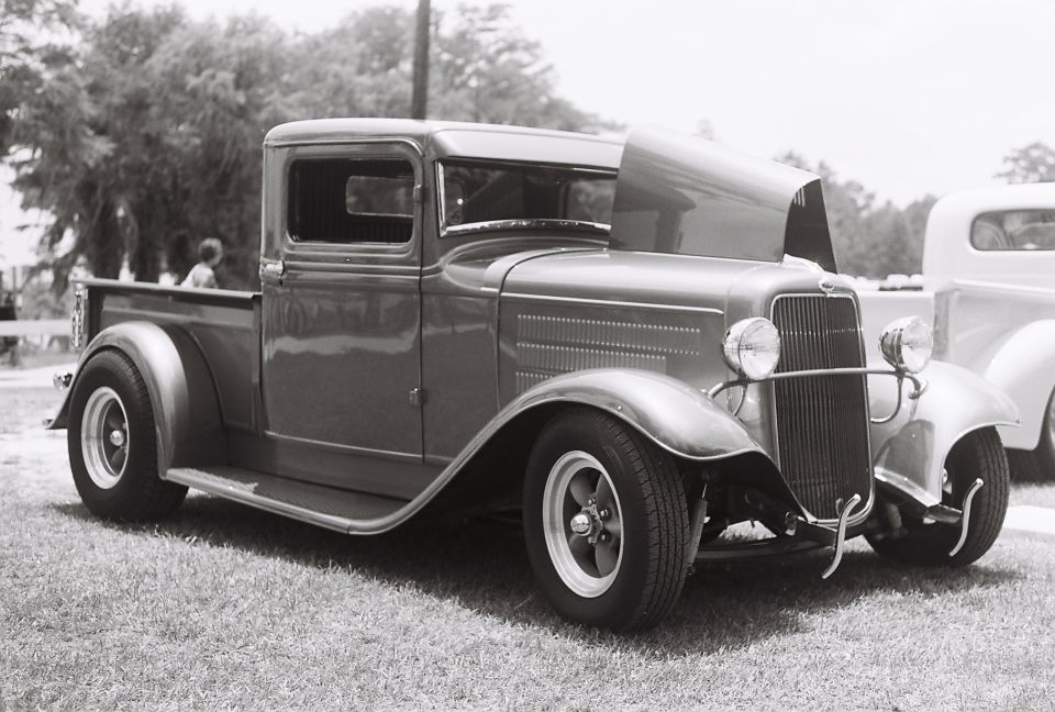 Modified 1932 Ford pickup truck with hood open, parked on grass. Another truck is visible to the right