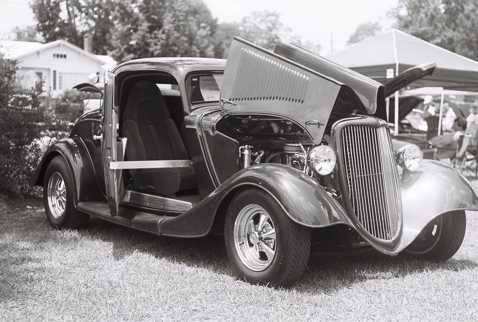 Dark hot rod coupe with hood open, showing engine. Chrome wheels, open door. Grille and headlights visible