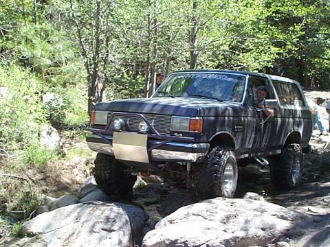 1989 Ford Bronco - This lifted Ford Bronco is shown off-roading on a rocky trail with trees in the background.