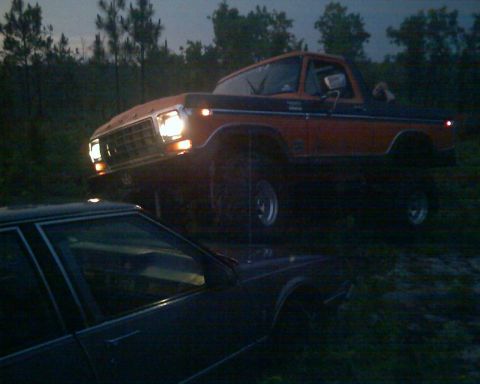 1979 Ford Bronco - A lifted 1979 Ford Bronco is seen in a wooded area at dusk, with its headlights illuminated.