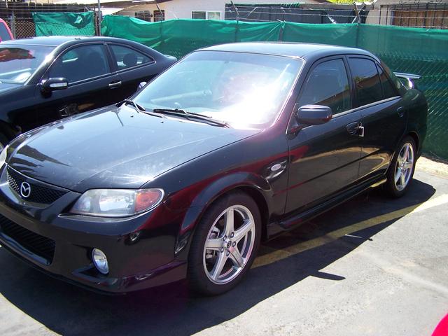 Black Mazda sedan, 3/4 front view, silver five-spoke wheels and rear wing, by a green fence
