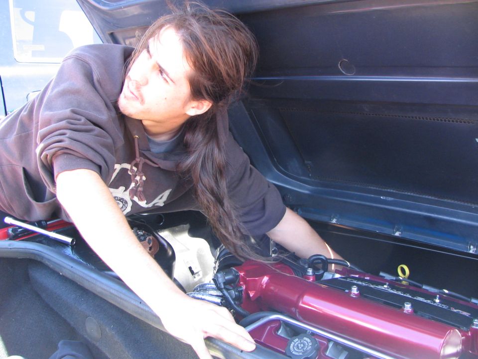 Person leaning into an engine bay, working on a red engine component. Supermotors.net logo visible