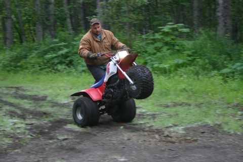 2003 Jeep TJ - A man rides a three-wheeled ATV, lifting the front wheels off the ground in a wheelie on a dirt path surrounded by trees.