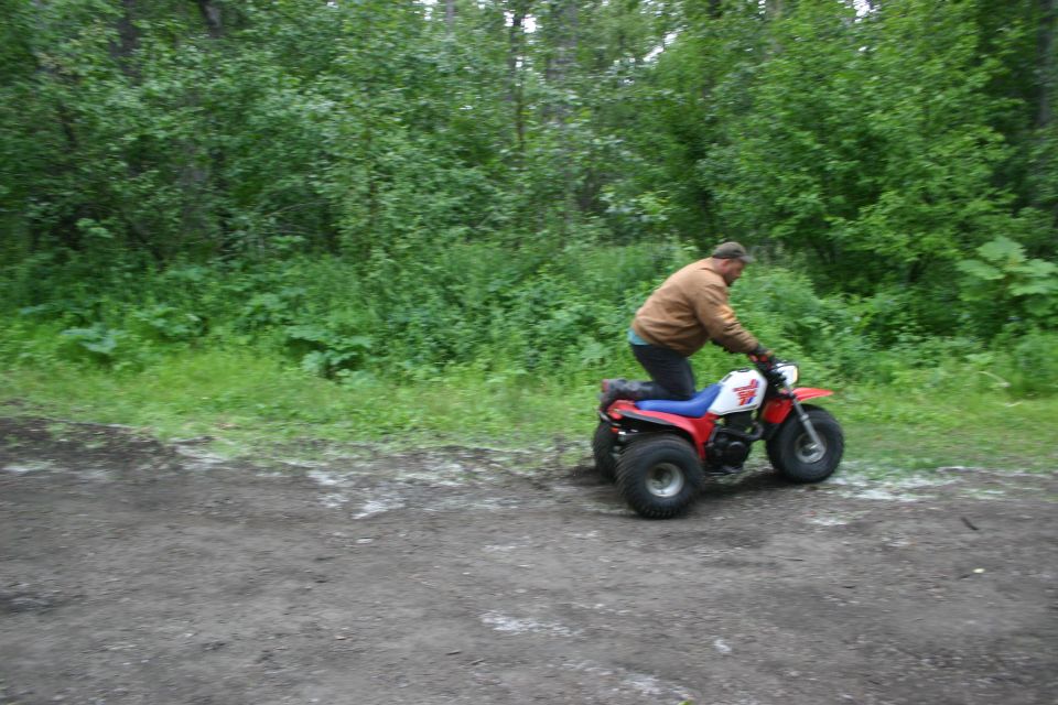 Man riding a red, white, and blue three-wheeled ATV on a dirt path with trees in the background