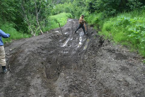 2003 Jeep TJ - Two people navigate a severely rutted and muddy trail in a wooded area.