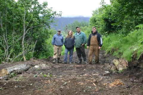 2003 Jeep TJ - A group of four individuals stand on a muddy trail, with felled logs and vegetation visible in the background.