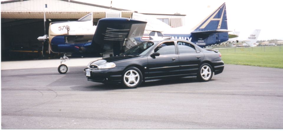 Black Ford Contour sedan with hood up on airfield apron, Navy aircraft parked behind