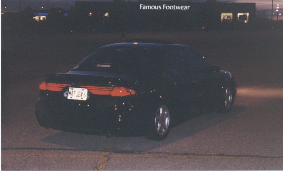 Dark sedan rear three-quarter at night in parking lot, Famous Footwear store lit behind