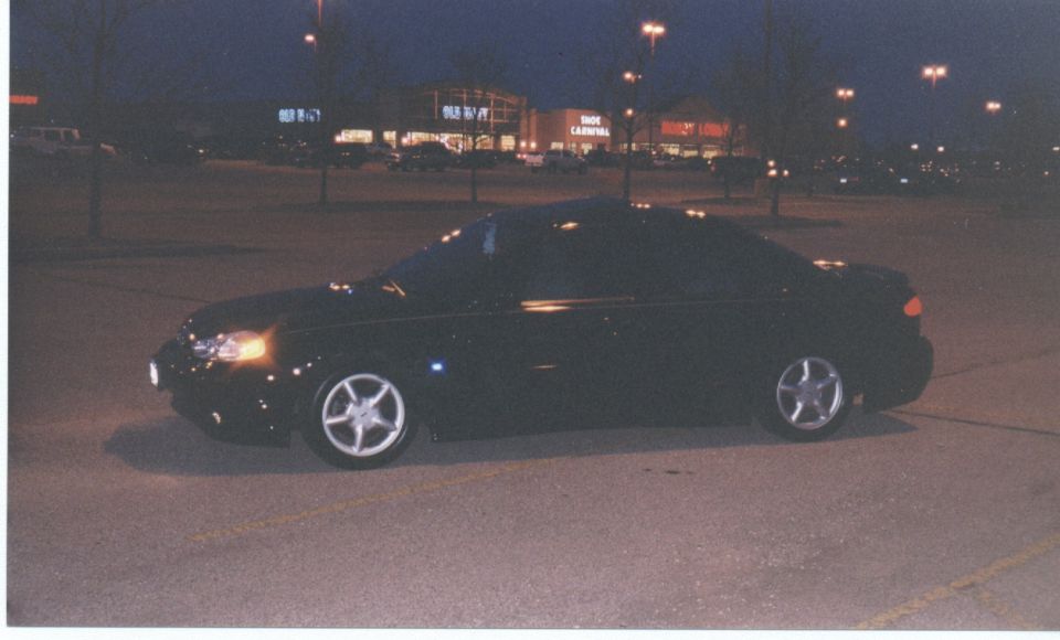 Black coupe side profile at night, headlights on, five-spoke wheels, lit storefronts behind