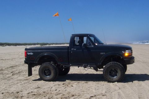 1994 Ford F-150 - A lifted black pickup truck sits on a sandy beach under a clear blue sky, with two orange flags flying from its bed.