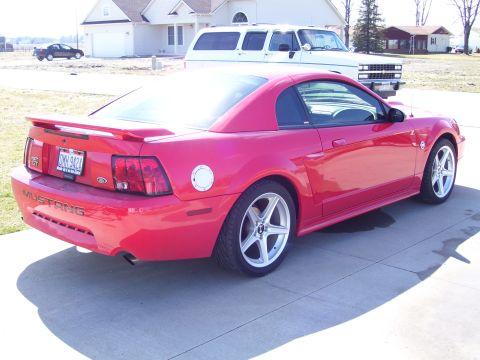 2004 Ford Mustang - A red 2004 Ford Mustang is parked in front of a house, with other vehicles visible in the background.