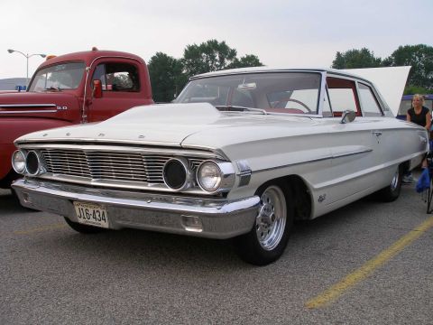 1998 Ford Crown Victoria - A white 1964 Ford Galaxie sedan, modified with a hood scoop, is parked next to a red truck.