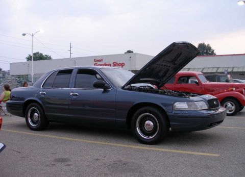 1998 Ford Crown Victoria - A dark blue sedan, possibly a Ford Crown Victoria, is parked with its hood open next to a red vintage car.