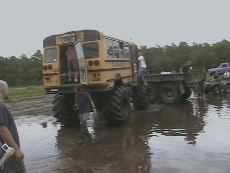 Big yellow school bus on huge tires in mud, with a flatbed truck and other vehicles nearby