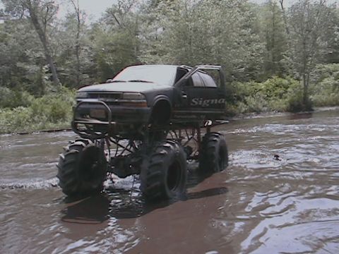 1976 Chevrolet Blazer - A heavily modified black truck with oversized tires navigates a shallow, muddy waterway.