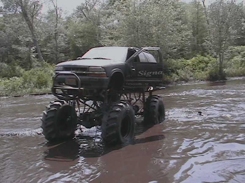 Black monster truck with large tires driving through muddy water, trees in background