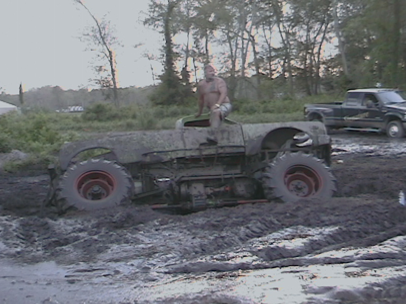 Mud-covered custom vehicle stuck in mud with a man sitting on it; black pickup truck in background