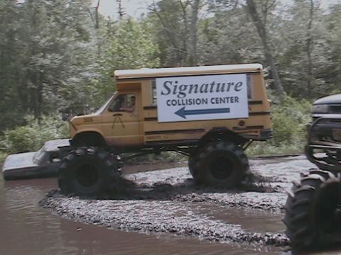 1976 Chevrolet Blazer - A heavily modified school bus with oversized tires navigates a muddy terrain, displaying 'Signature Collision Center' signage.