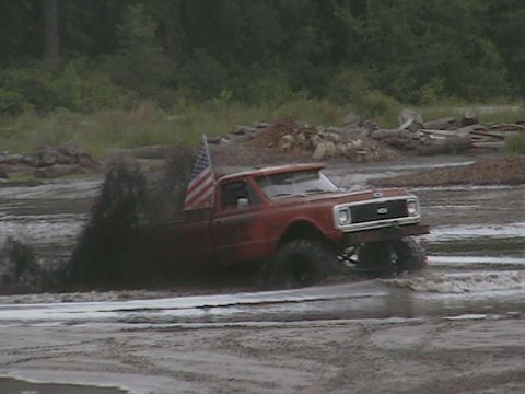 1976 Chevrolet Blazer - A red pickup truck with an American flag drives through a mud pit, kicking up a large spray.