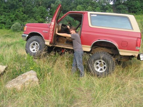 1988 Ford Bronco - A man leans into the open driver's door of a red and tan Ford Bronco parked on a grassy incline.