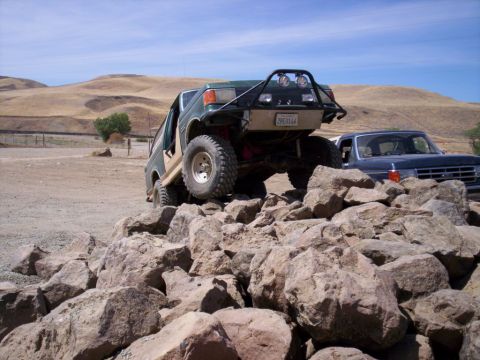 1989 Ford Bronco - A modified green SUV navigates a rocky obstacle course with a blue pickup truck in the background.