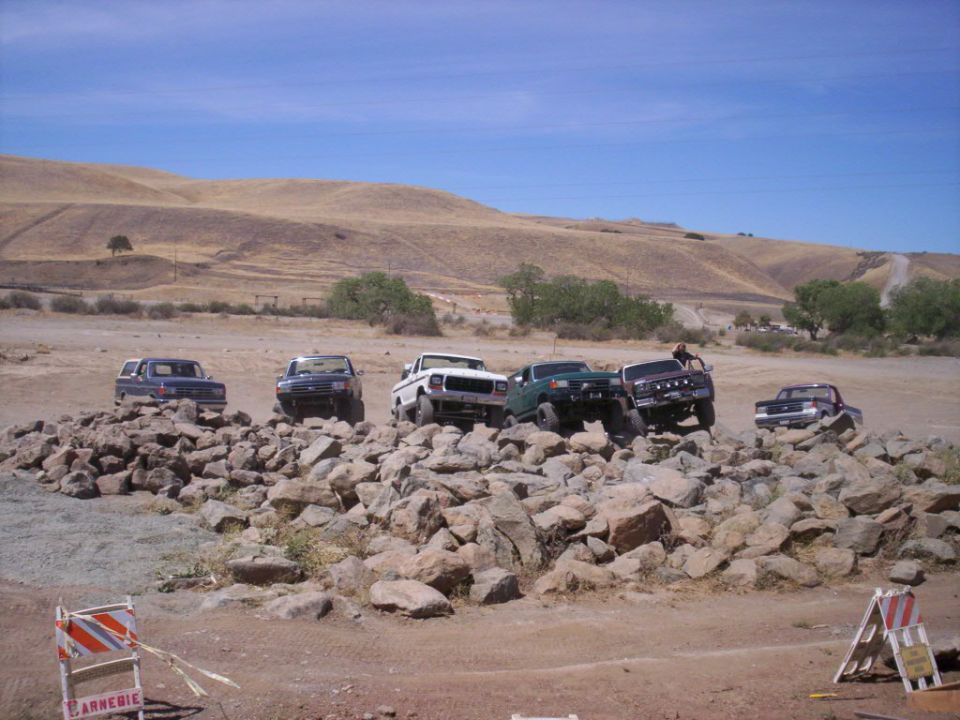 Several SUVs and trucks, including a blue Bronco-like vehicle and a white pickup, navigate a rocky obstacle course under a clear sky
