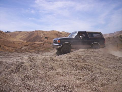 1989 Ford Bronco - A dark blue 1989 Ford Bronco with a tan top navigates a dusty, gravelly incline under a partly cloudy sky.