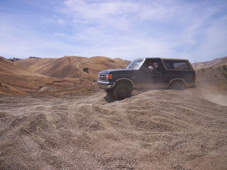 Dark blue 1989 Ford Bronco with tan top driving on a gravel hill