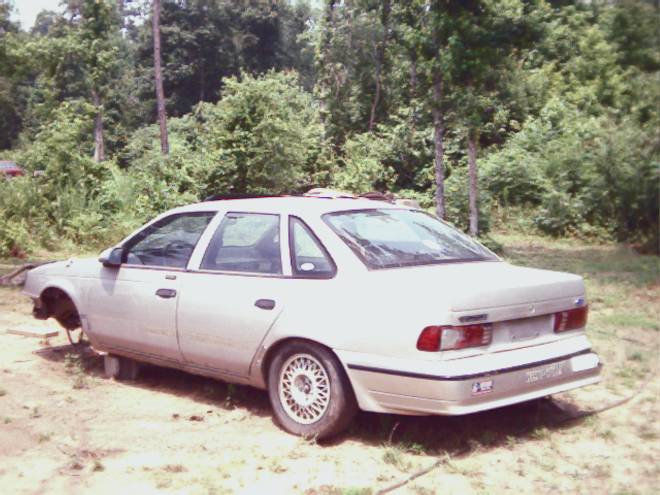 Silver Ford Taurus sedan in a grassy field, rear three-quarter view, front end resting on the ground