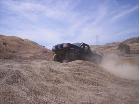1989 Ford Bronco - A dark pickup truck navigates a dusty, gravelly terrain under a partly cloudy sky.