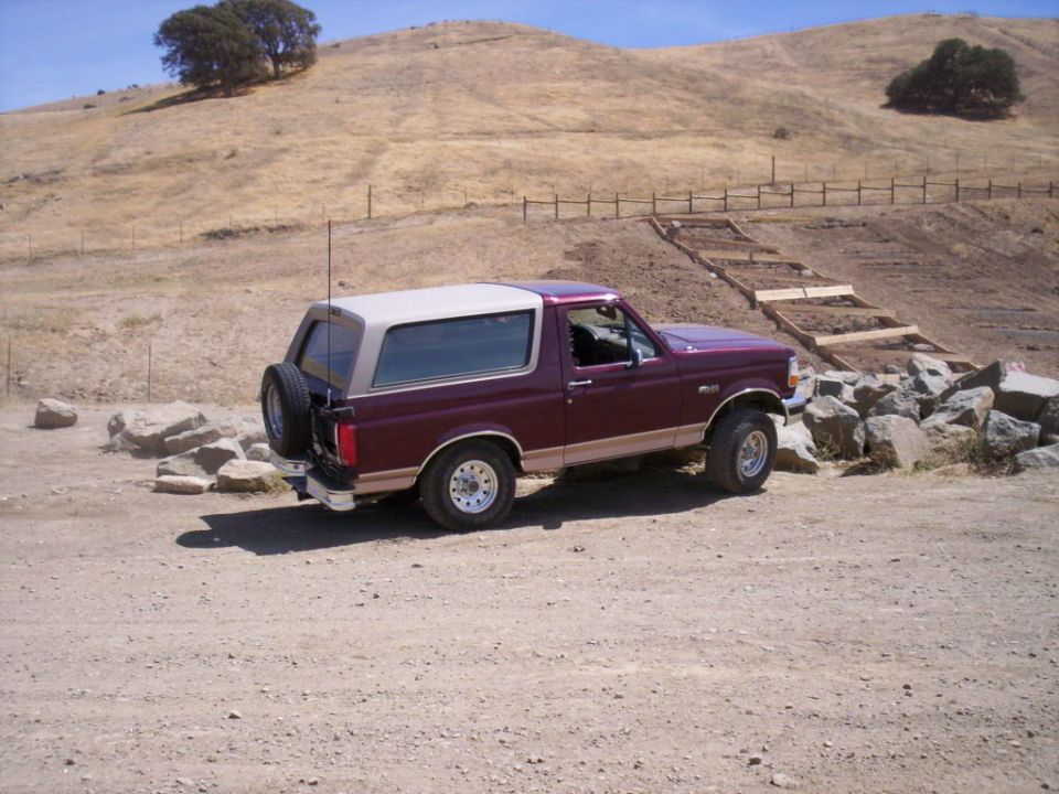 Maroon Ford Bronco with tan top parked on dirt road, spare tire on back. Hillside with trees and fence in background