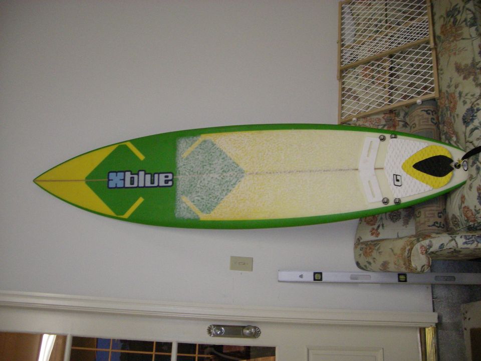 Green and yellow xblue surfboard leaning against an interior wall beside a floral couch
