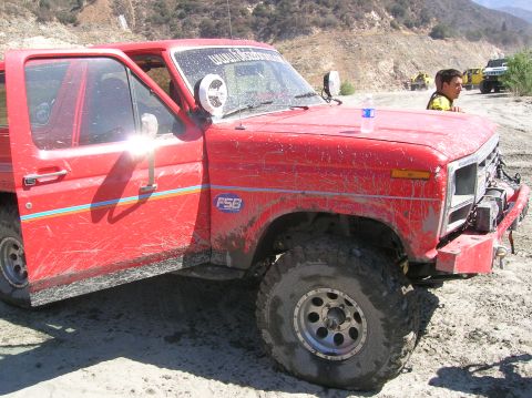 1985 Ford Bronco - A muddy red Ford Bronco sits off-road with its door ajar, while other vehicles are visible in the background.