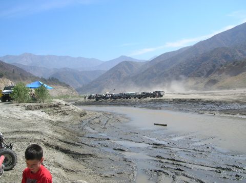 1985 Ford Bronco - A line of off-road vehicles are parked on a dirt road beside a muddy stream, with a mountainous landscape behind them.
