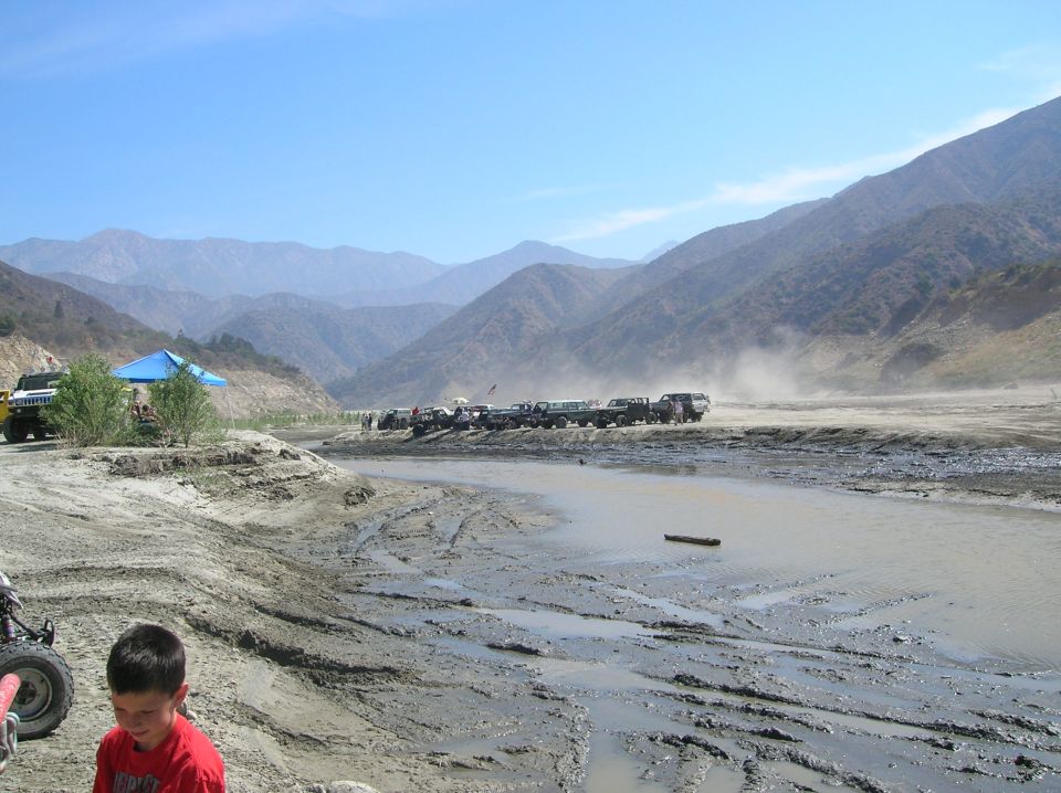 Off-road vehicles parked on dirt road by muddy stream, mountains in background