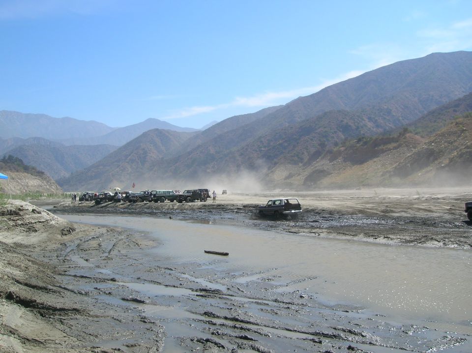 Several off-road vehicles parked and driving in a dusty, muddy riverbed with mountains in the background