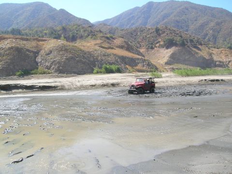 1985 Ford Bronco - A red off-road vehicle navigates a muddy terrain with arid hills under a clear blue sky.